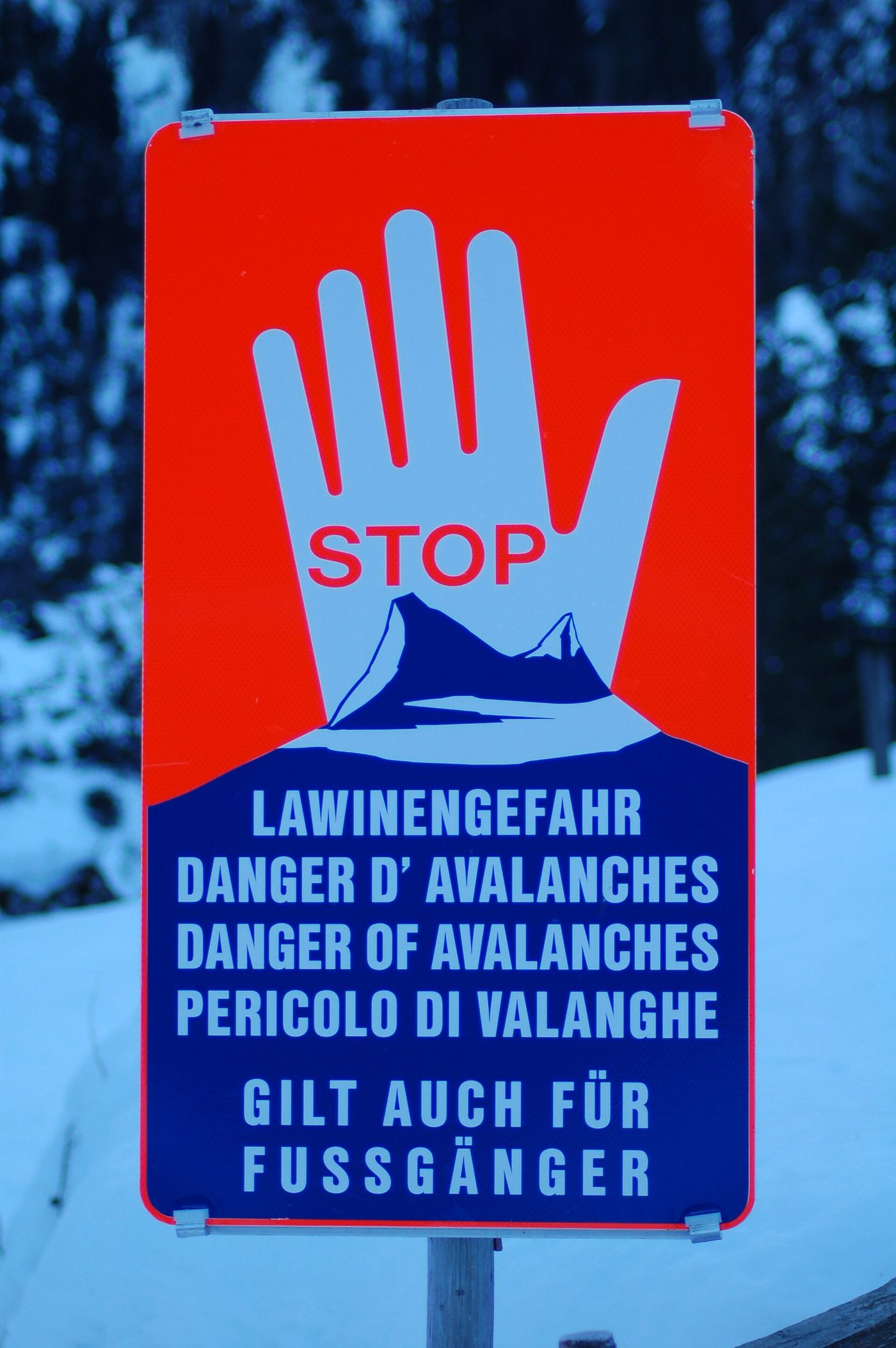 A red and blue STOP avalanche danger warning sign in the Austrian Alps, printed in four languages