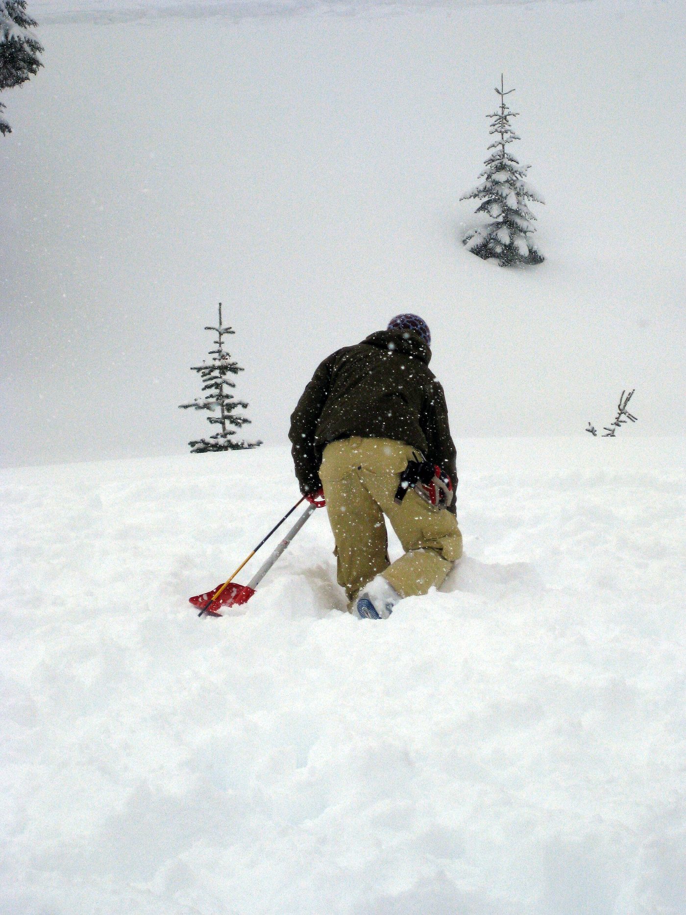 A skier kneeling in falling snow practicing a companion rescue with a probe and shovel