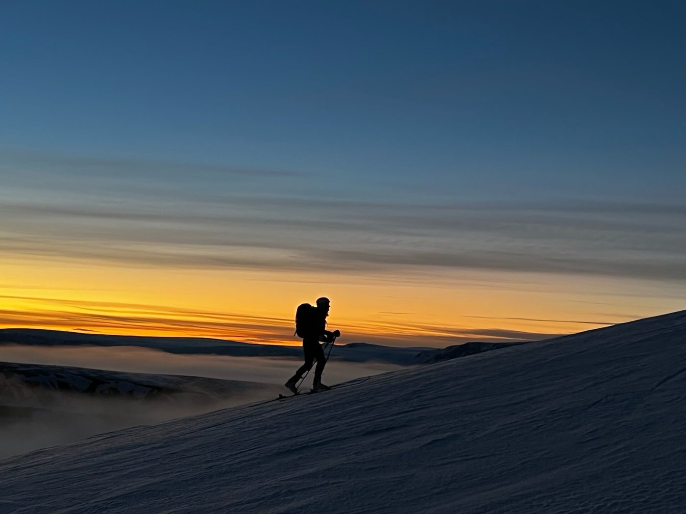 A backcountry skier silhouetted against an orange sunrise while climbing a snow ridge on skins