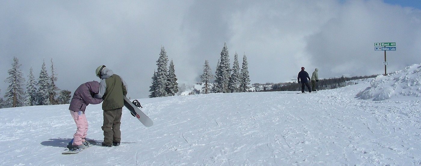 Two snowboarders pause near trail signs for Hidden Lake at Powder Mountain, Utah, with clouds rolling over the ridge