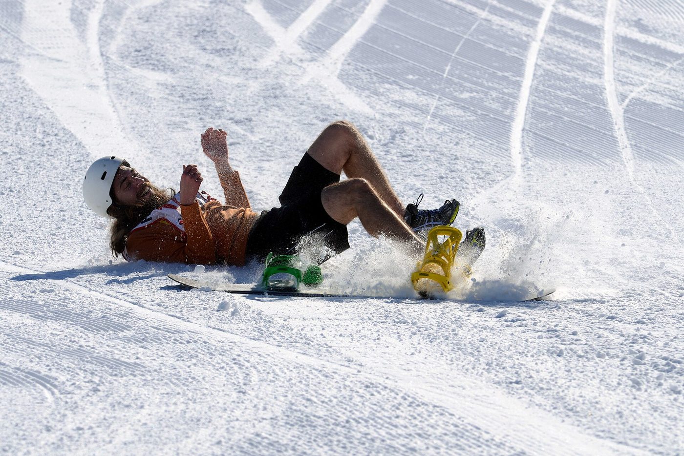 A snowboarder falling backward onto a groomed slope, arms raised