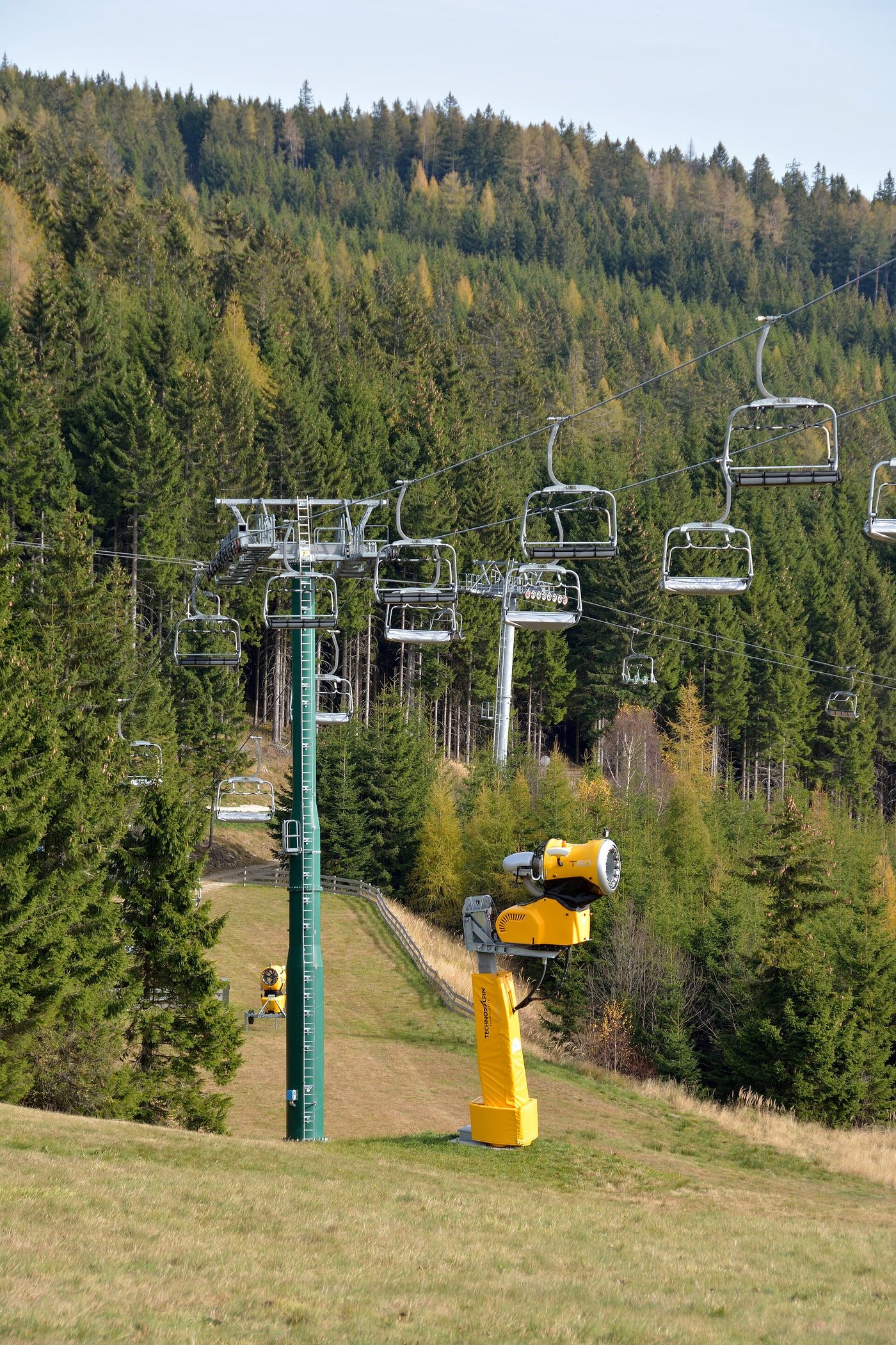 A snow gun beside an empty chairlift on a green autumn slope, the time of year many skiers first ask when to buy a ski pass